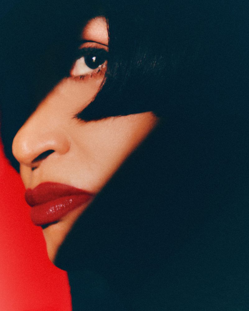 Extreme close-up beauty portrait of model Farida Khelfa with bold red lipstick and black bangs against a red backdrop.