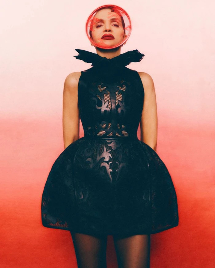 Model Farida Khelfa wearing a black structured dress and red circular face accessory against a red gradient background.