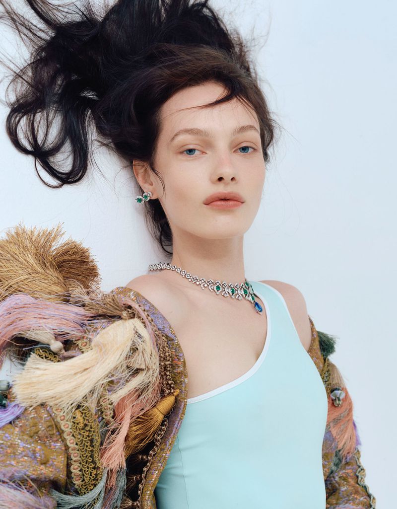 Close-up of a young woman lying down wearing a turquoise tank top, a colorful fringed jacket, and jewelry against a white background.