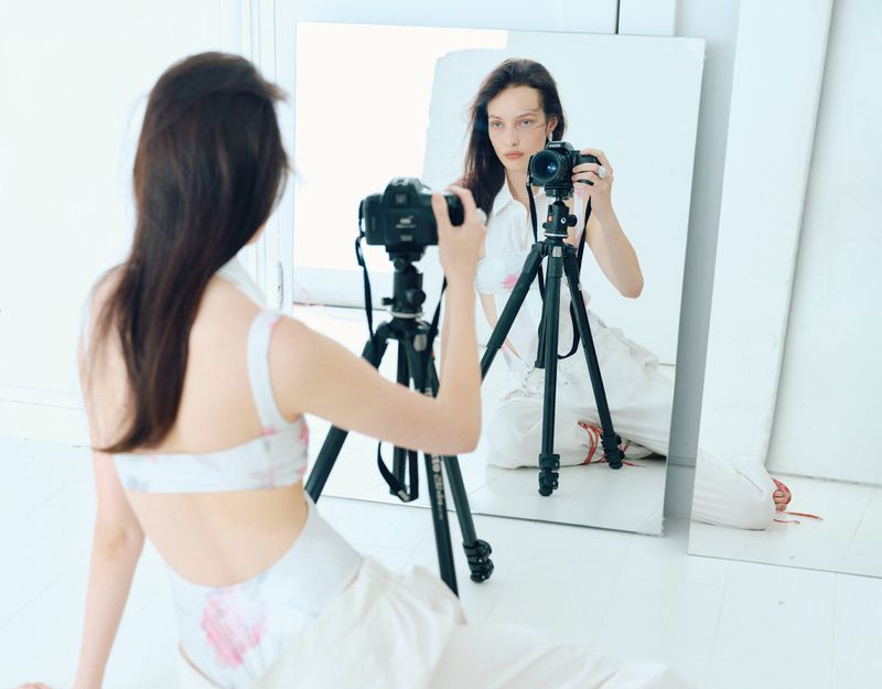 Woman photographs her reflection in a mirror using a DSLR camera on a tripod in a bright studio, wearing a halter top.