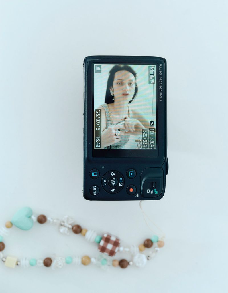 A digital camera displays a portrait of a woman with dark hair and earrings on its screen, while a beaded accessory lies nearby on a white surface.