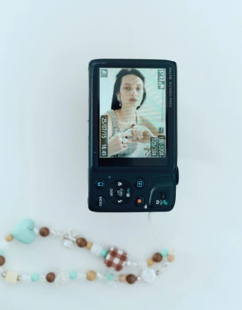 A digital camera displays a portrait of a woman with dark hair and earrings on its screen, while a beaded accessory lies nearby on a white surface.