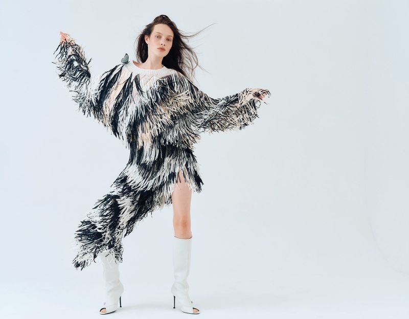 A stylish adult female model in a fringed black and white dress, striking a dynamic pose with white boots in a white photo studio.