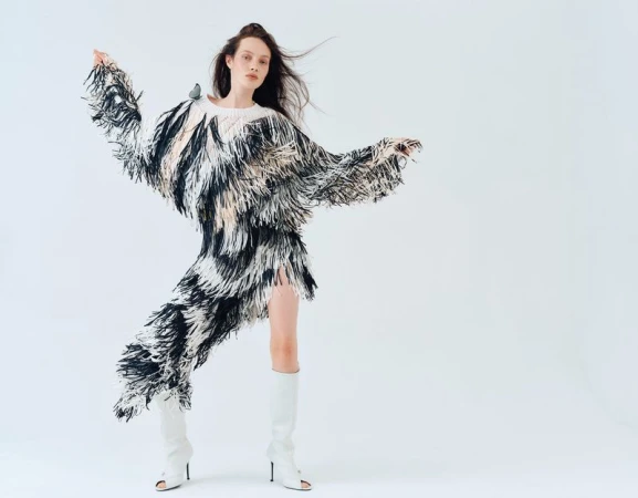 A stylish adult female model in a fringed black and white dress, striking a dynamic pose with white boots in a white photo studio.