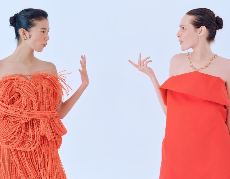 Two women in strapless orange gowns face each other in a studio, one in a spaghetti-like dress, the other in a sleek design.