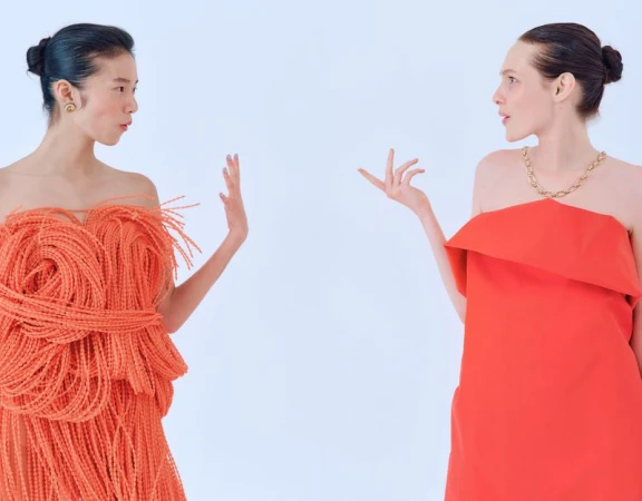 Two women in strapless orange gowns face each other in a studio, one in a spaghetti-like dress, the other in a sleek design.