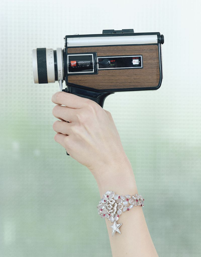 Adult female holds a vintage Super 8 camera with a silver and gemstone floral bracelet in a softly-lit indoor environment.