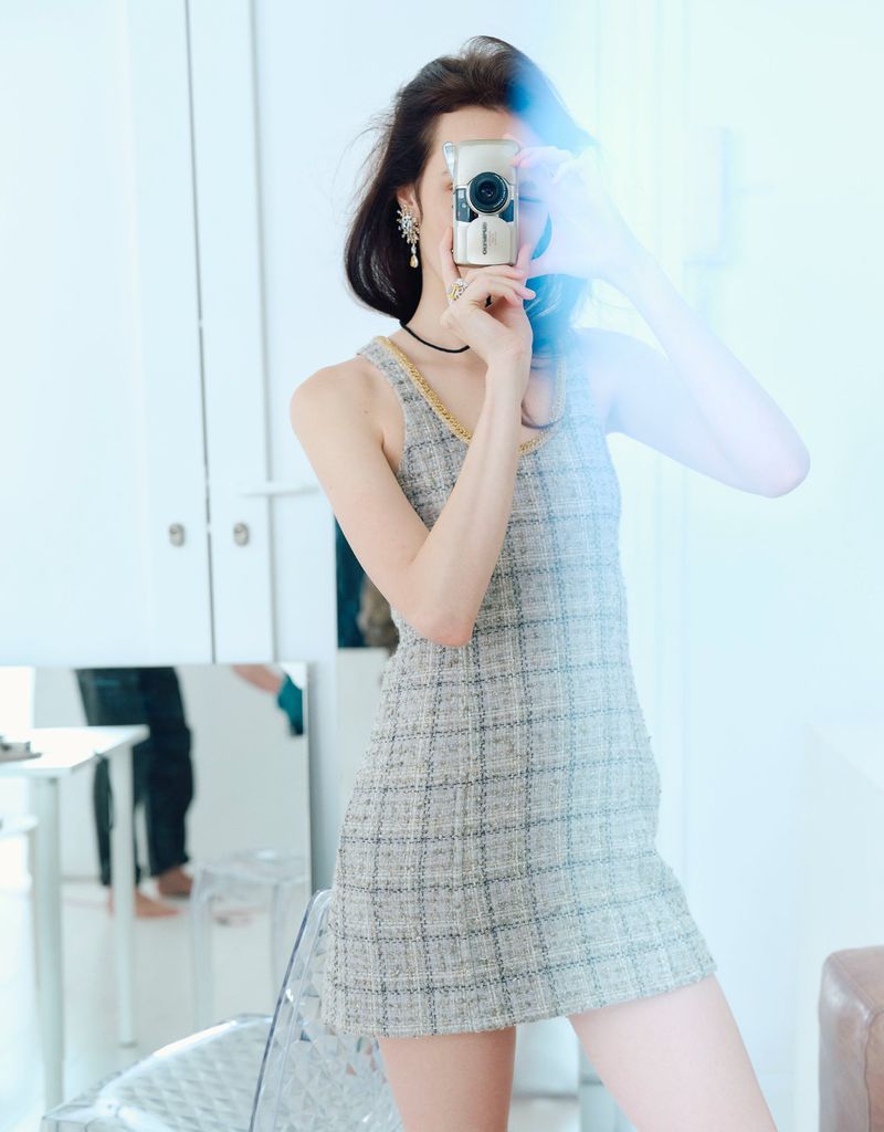 A woman in a gray dress holds a camera up to her face in an indoor setting with natural light and a visible mirror reflection.