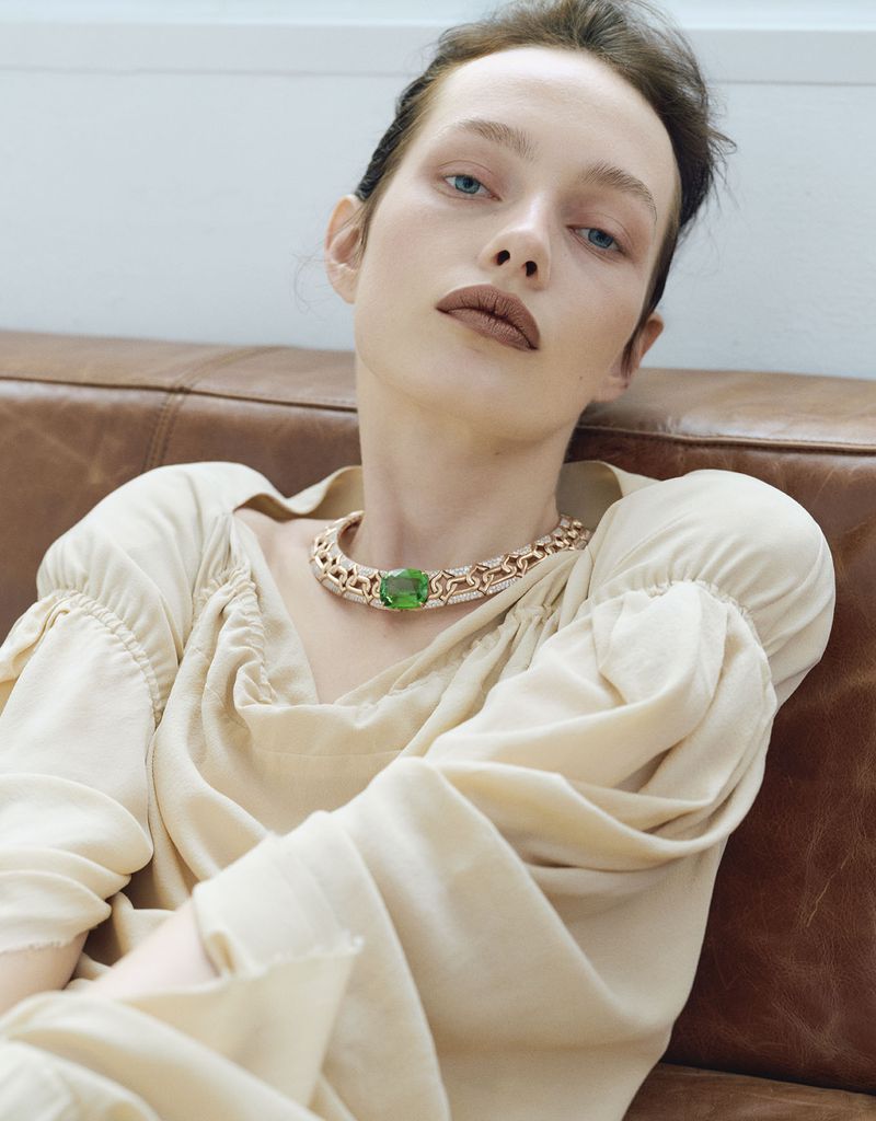 Close-up portrait of a woman with a necklace featuring a large green gemstone, lounging on a brown leather couch in natural light.