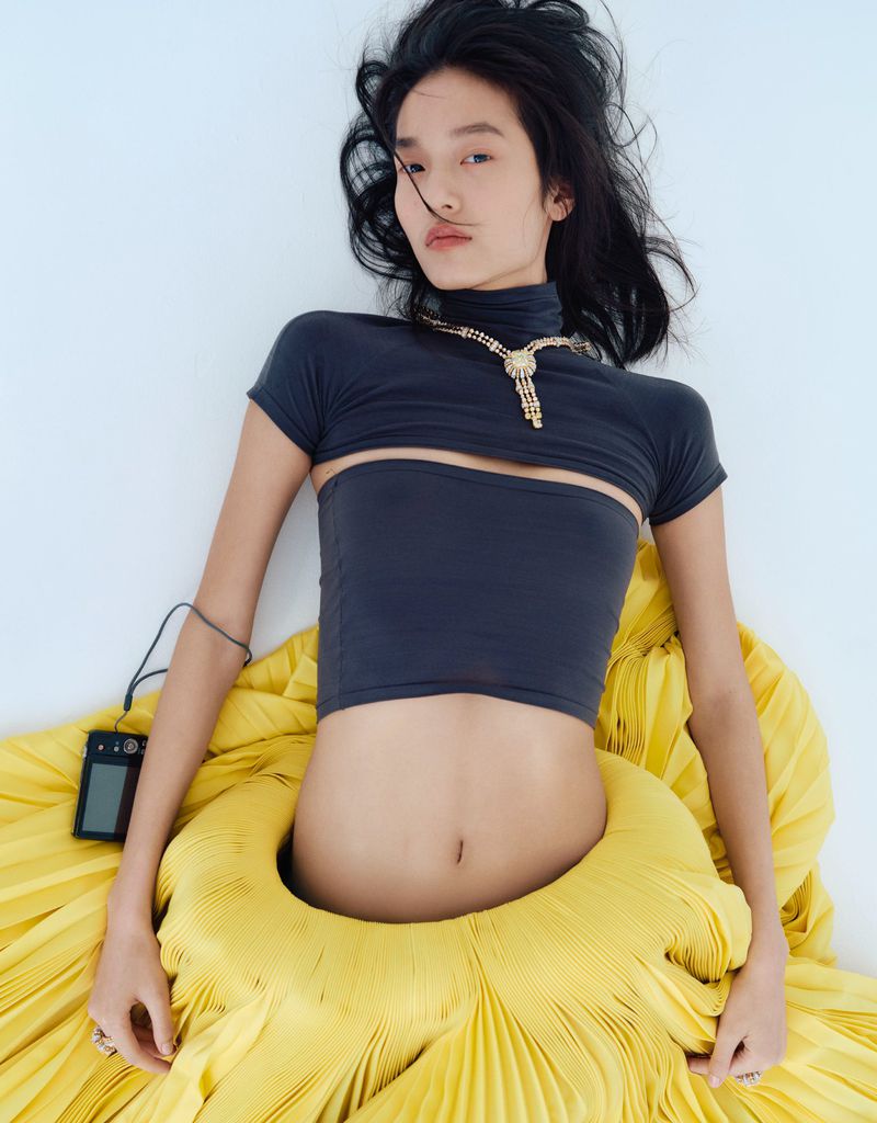 Asian woman lying on a white background wearing a gray top and yellow pleated skirt. She has a gold necklace and carries a device.