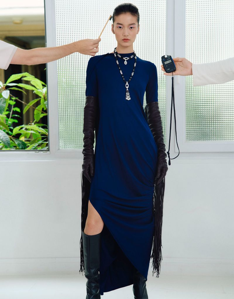 A slender woman in a stylish dark blue gown poses in front of a bright window, while two people off-camera hold props near her.