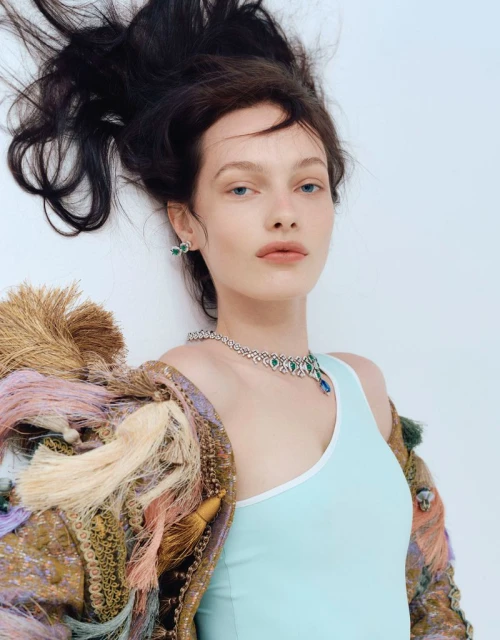 Model with dark hair wearing a light blue top and embellished fringe jacket with a diamond necklace against a white background