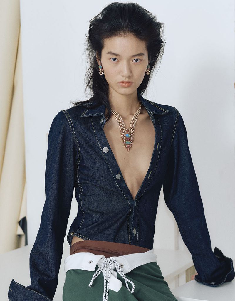 Model wearing a dark denim shirt and layered pendant necklace against a plain white studio backdrop.