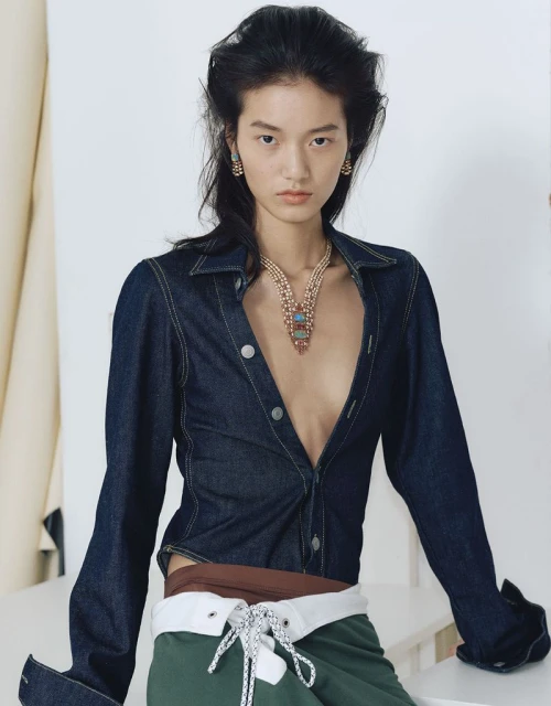 Model wearing a dark denim shirt and layered pendant necklace against a plain white studio backdrop.