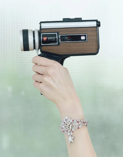 Hand holding a vintage Super 8 camera wearing an ornate diamond and pink gemstone bracelet with a star charm against a green backdrop.