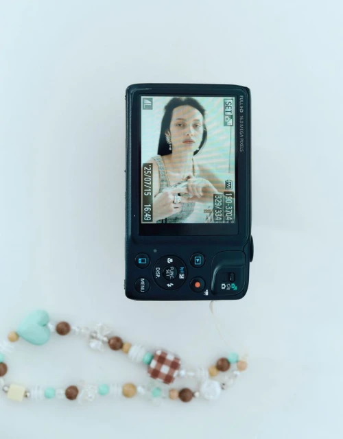 Compact digital camera displaying a model portrait on its LCD screen with a beaded strap resting on a white surface.