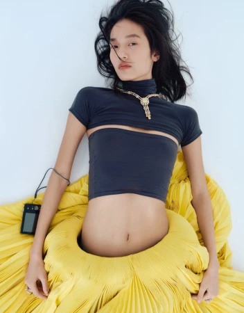 Model Qiuyue Li lying down in a charcoal crop top and large yellow pleated skirt against a white studio background.