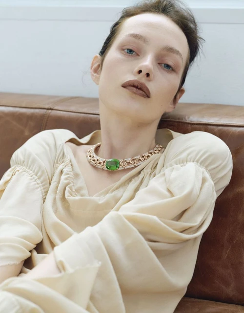 Model Clara Denison in a cream puff sleeve top and green gemstone choker necklace reclining against a leather sofa.