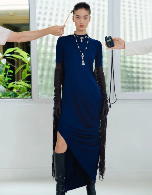 Model posing in a navy blue maxi dress, long fringe gloves, and black boots while crew hands hold a light meter and brush.