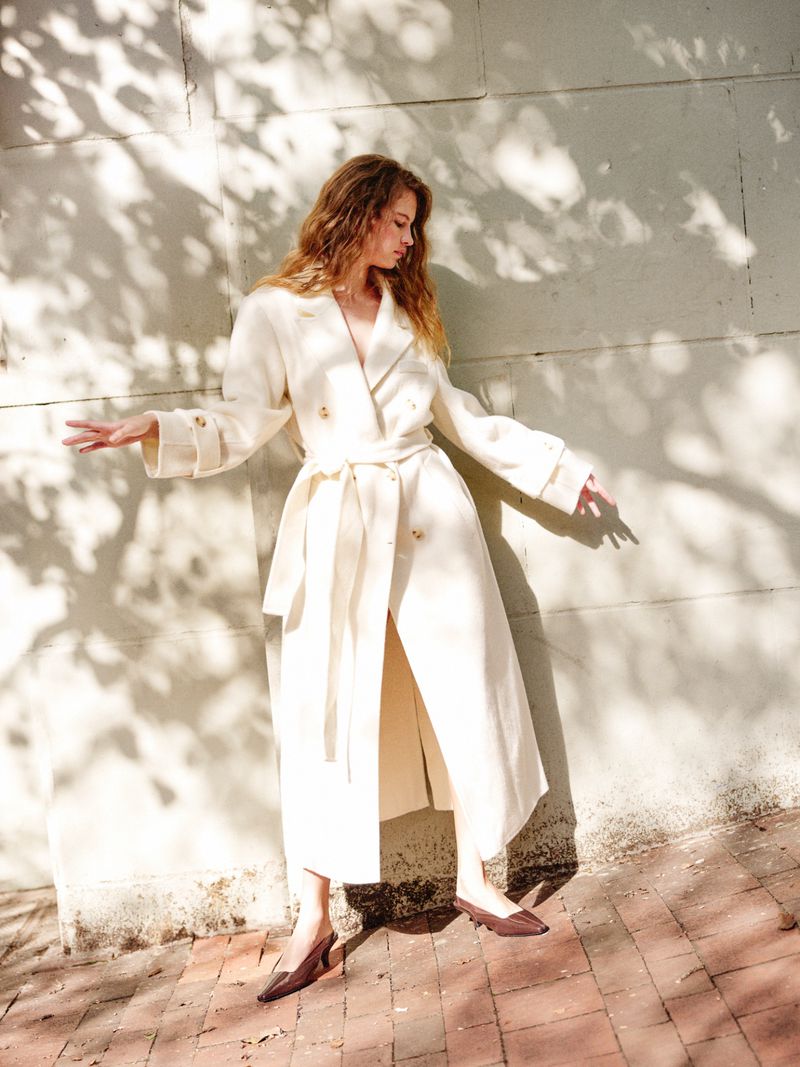 Fashionable woman in trench coat poses outdoors in sunlight with long brown hair, showcasing elegant brown pumps and stylish outfit.