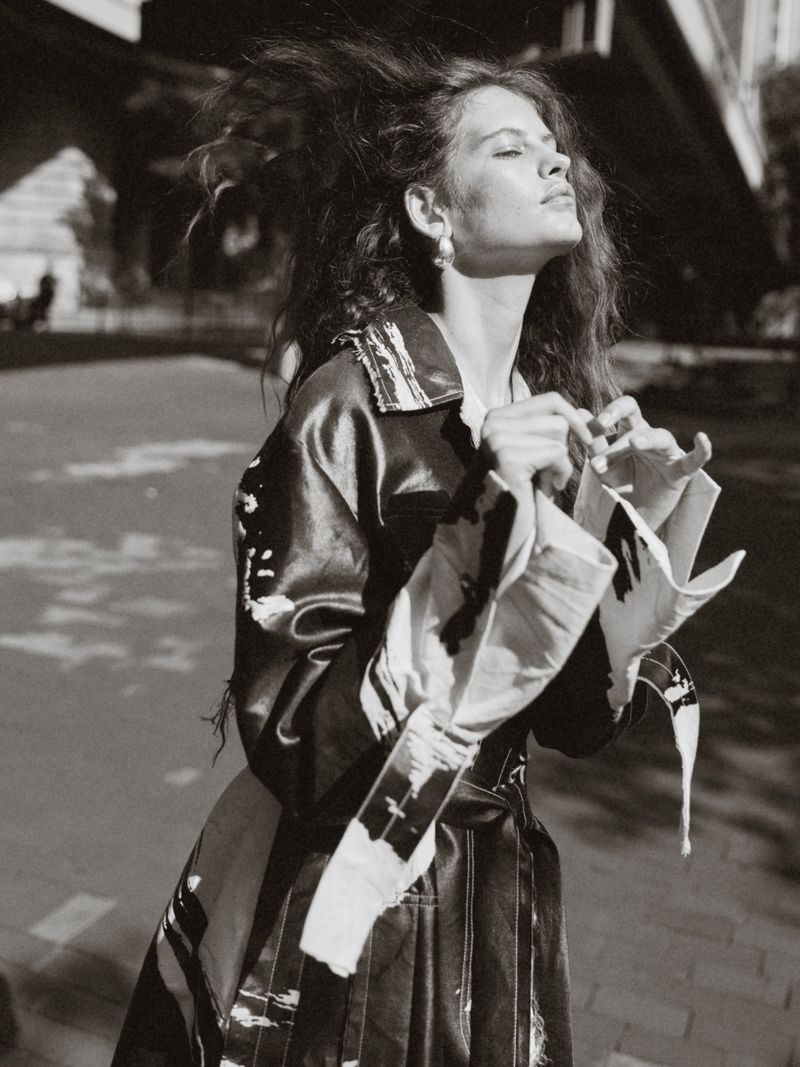 Monochrome outdoor shot of a woman with tousled hair in an abstract-print coat, standing on a paved surface near an urban structure.