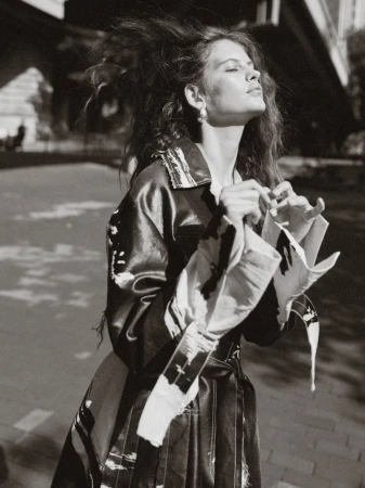 Monochrome outdoor shot of a woman with tousled hair in an abstract-print coat, standing on a paved surface near an urban structure.