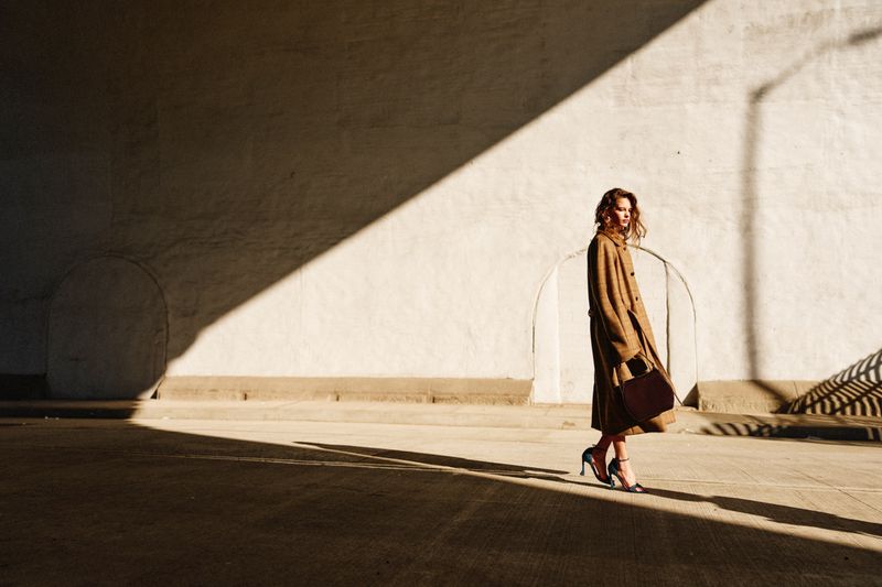 A woman in a tan coat and blue heels walks across a sunlit concrete expanse under a shadowed wall with an arched doorway.