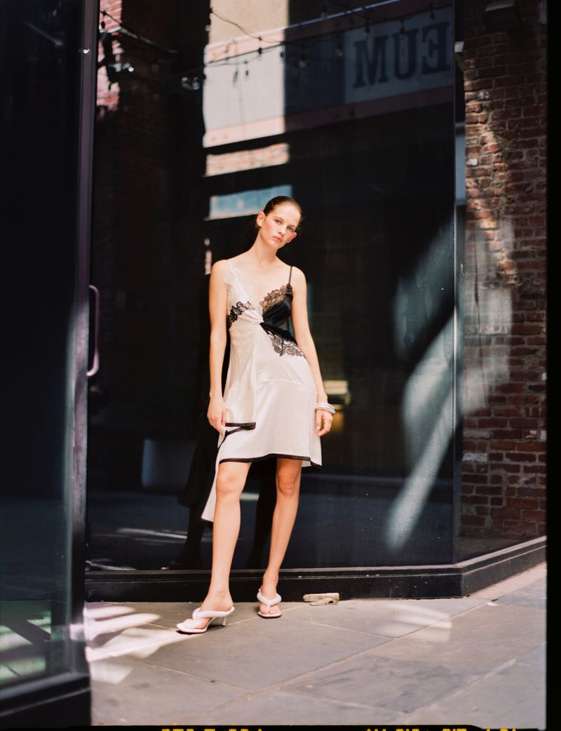 Woman in a satin dress with lace detail stands in a doorway on a city street. Street scene with brick buildings and a shadowed storefront.