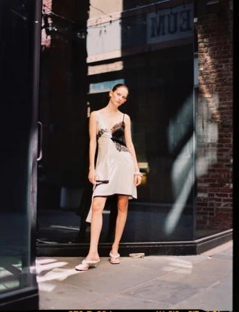 Woman in a satin dress with lace detail stands in a doorway on a city street. Street scene with brick buildings and a shadowed storefront.