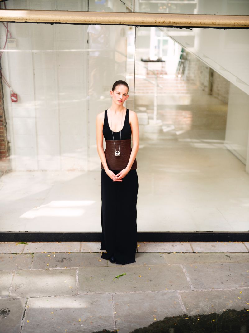 Full-length portrait of a woman in a black dress with a pendant necklace, standing in front of a large window and stone pavement.