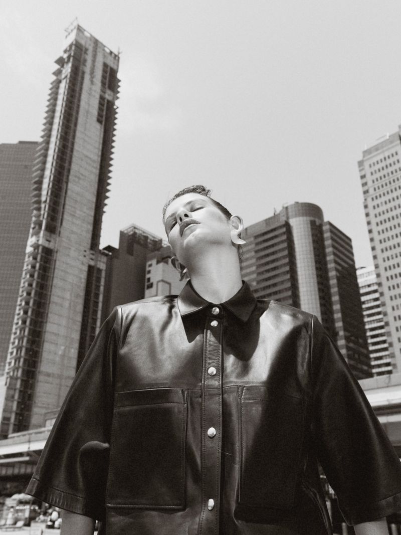 Monochrome portrait of a woman in a leather jacket against a backdrop of tall city buildings, showcasing urban fashion and style.