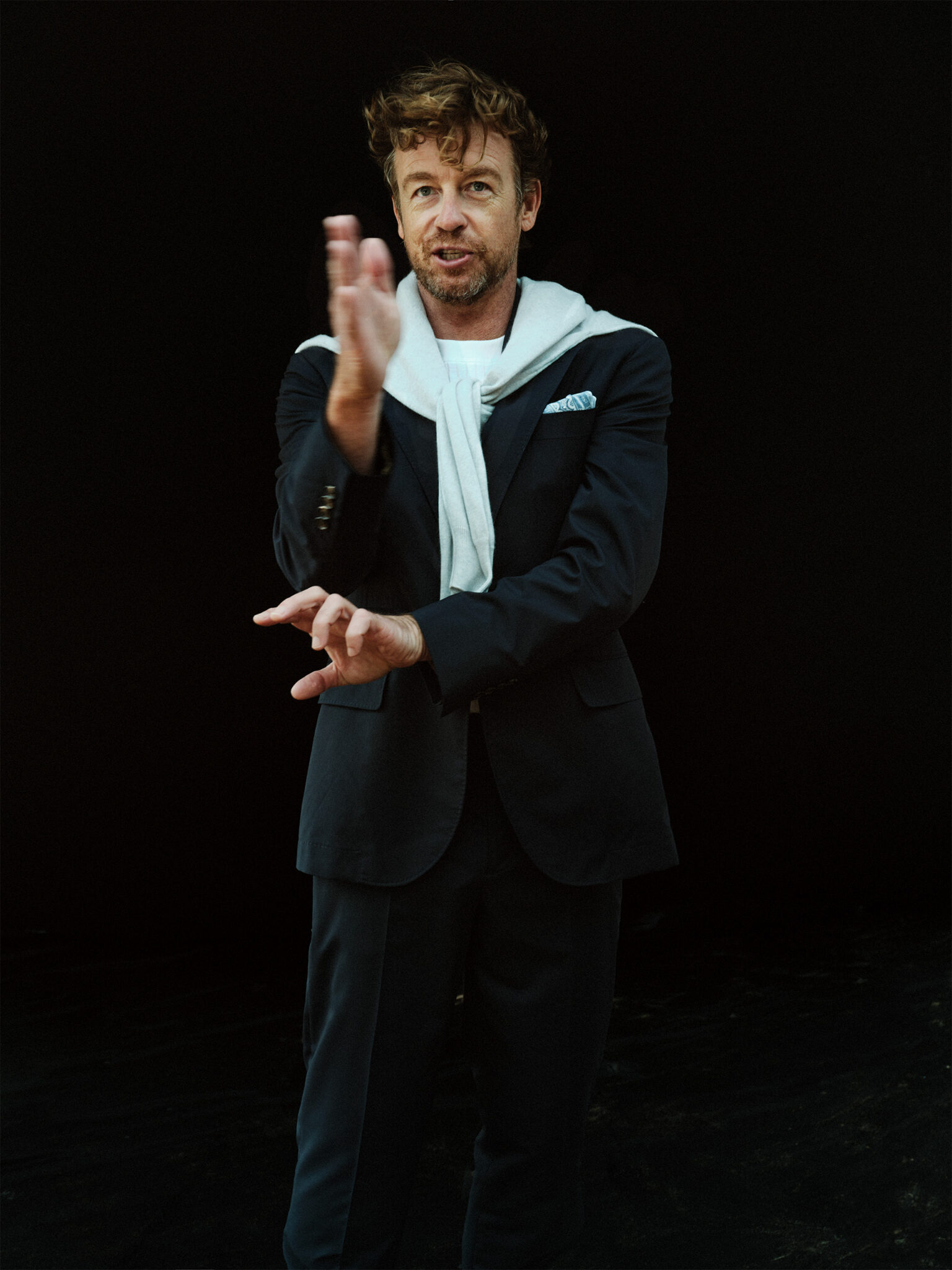 Portrait of adult male wearing a black suit with a white shirt and sweater, standing in front of a dark background, gesturing with hands.