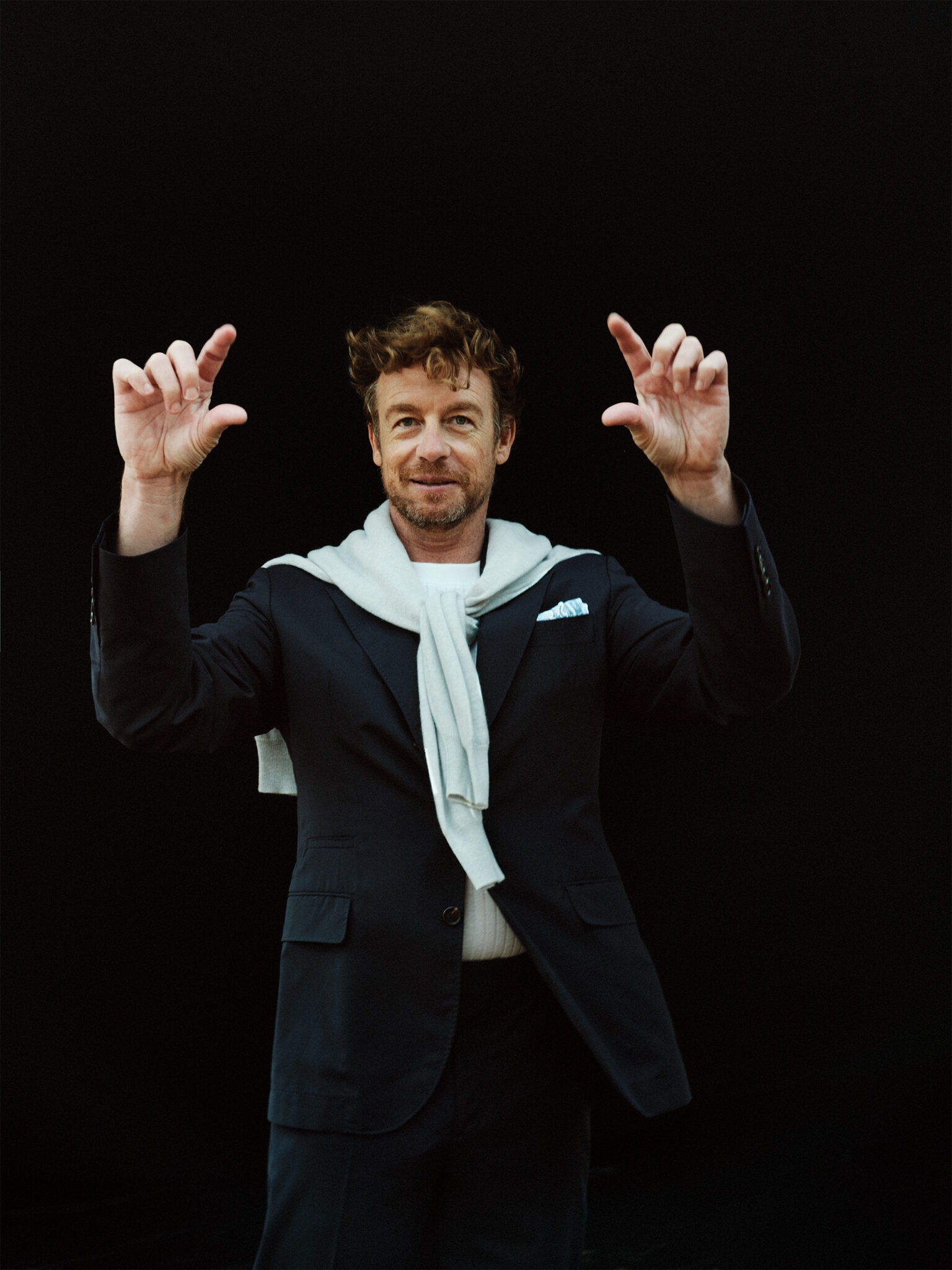 Portrait of an adult man in a dark suit and light scarf against a black backdrop, making a small gesture with his fingers.