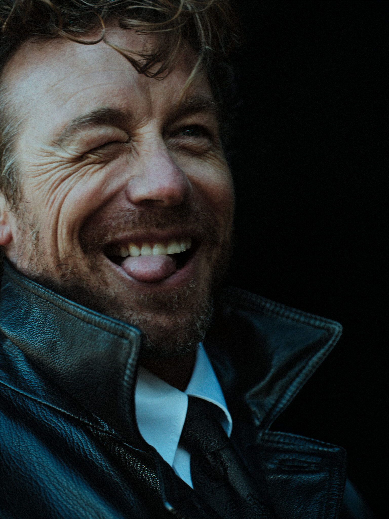 Portrait of a man with curly hair wearing a leather jacket, white shirt, and tie, smiling while sticking his tongue out and winking.