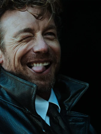 Portrait of a man with curly hair wearing a leather jacket, white shirt, and tie, smiling while sticking his tongue out and winking.
