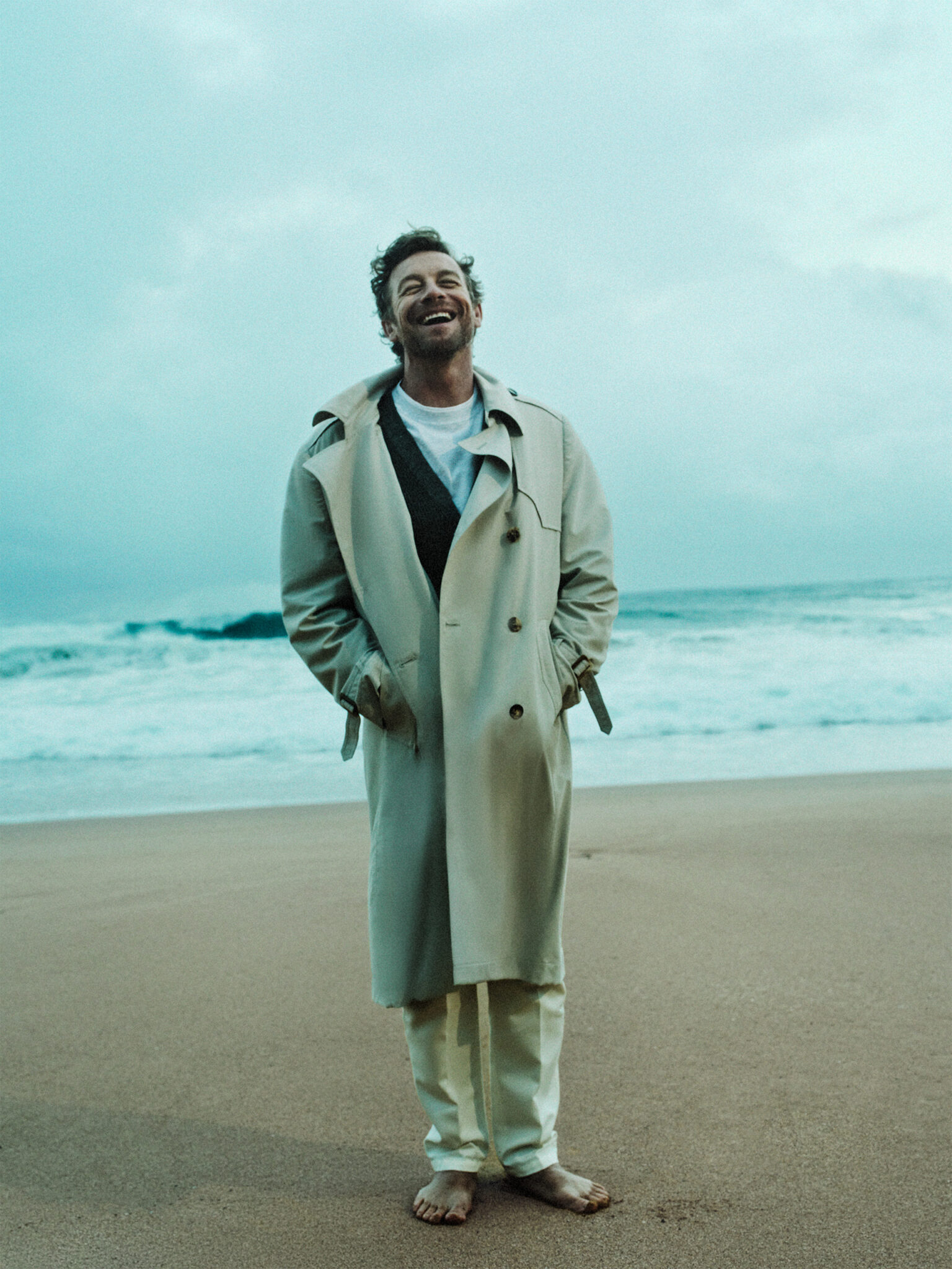 Adult male in a tan trench coat stands barefoot on a beach with an ocean background, smiling and looking upwards in a portrait-style shot.