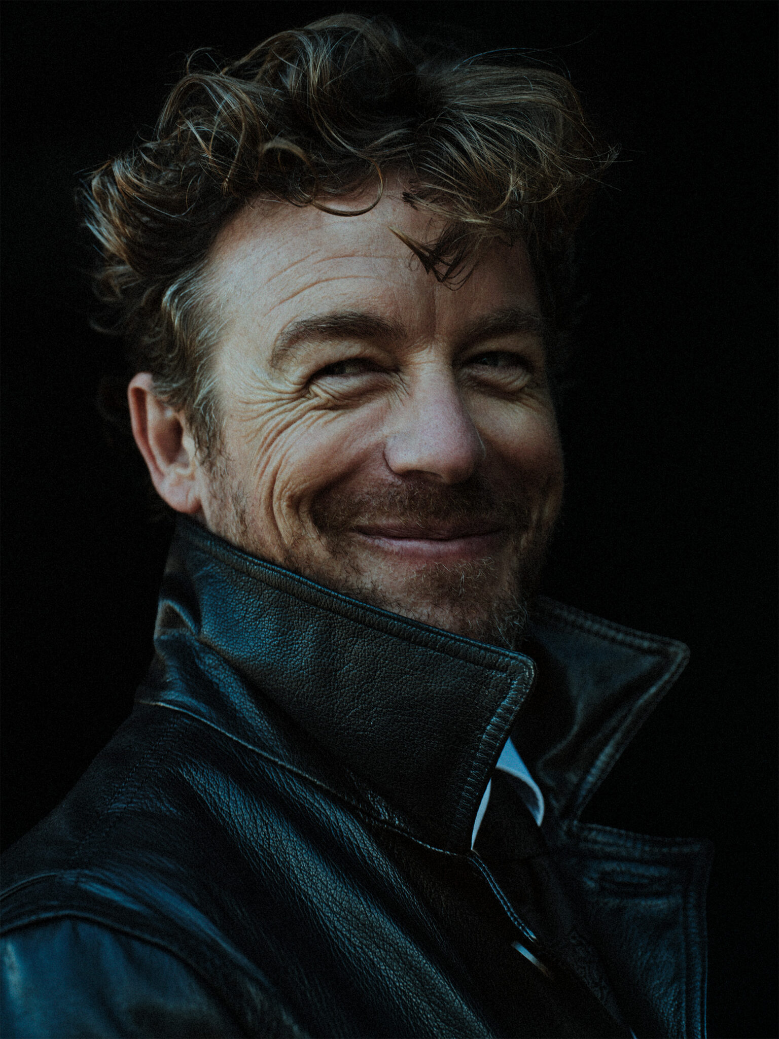 Portrait of a man with curly hair, a slight smile, and wearing a black leather jacket against a dark background.