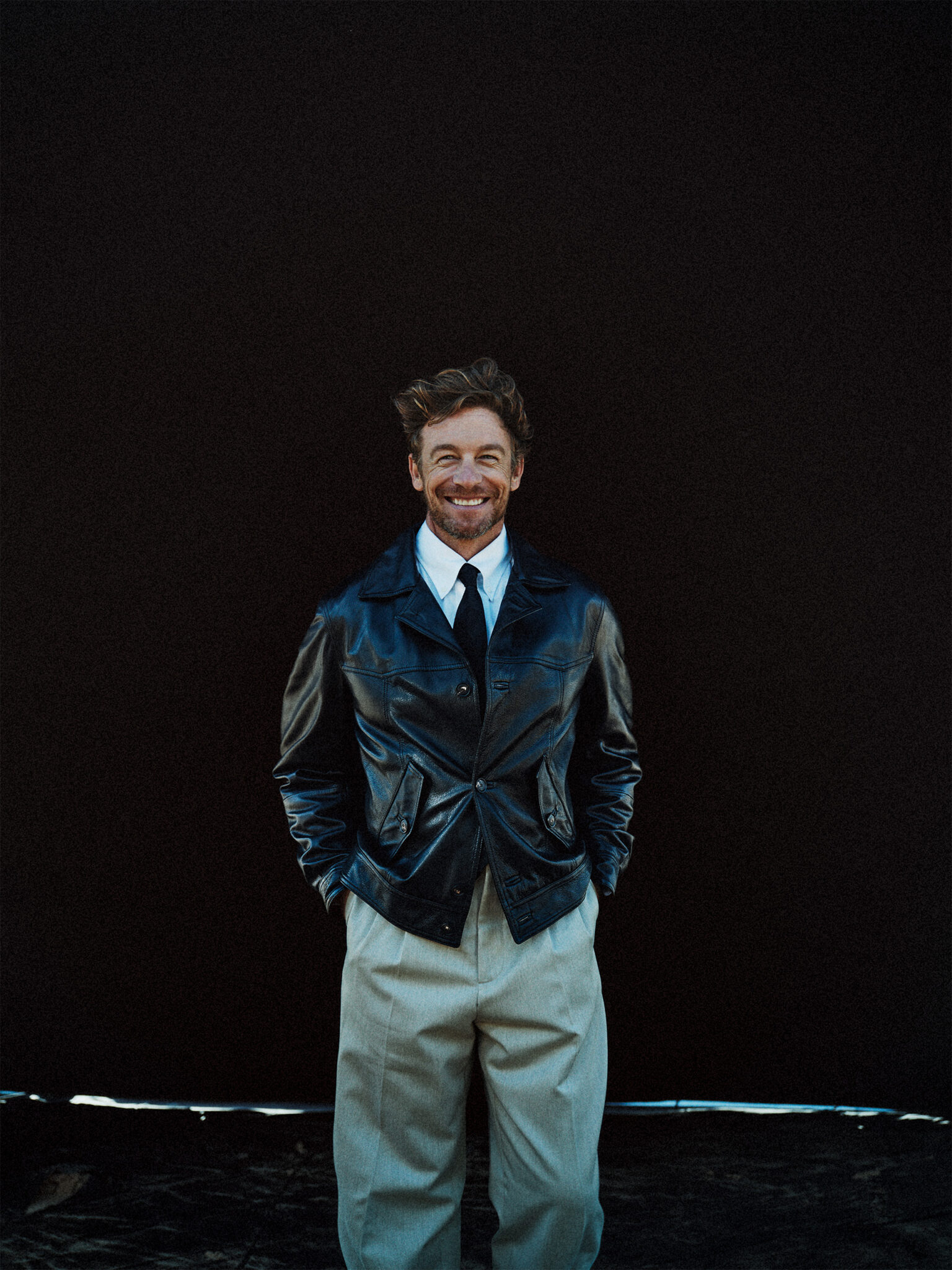 Smiling man in a leather jacket stands against a dark background with hands in pockets, captured in a studio portrait.