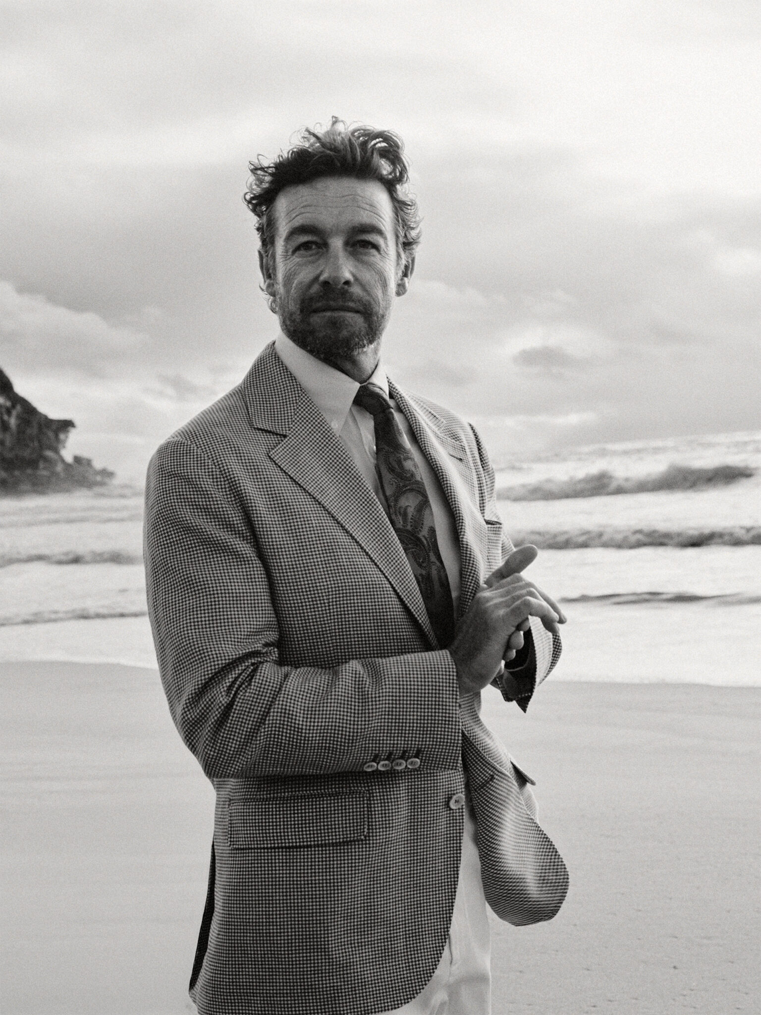 Monochrome portrait of a handsome man in a houndstooth blazer standing on a sandy beach with waves and cliffs in the background.