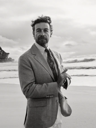 Monochrome portrait of a handsome man in a houndstooth blazer standing on a sandy beach with waves and cliffs in the background.