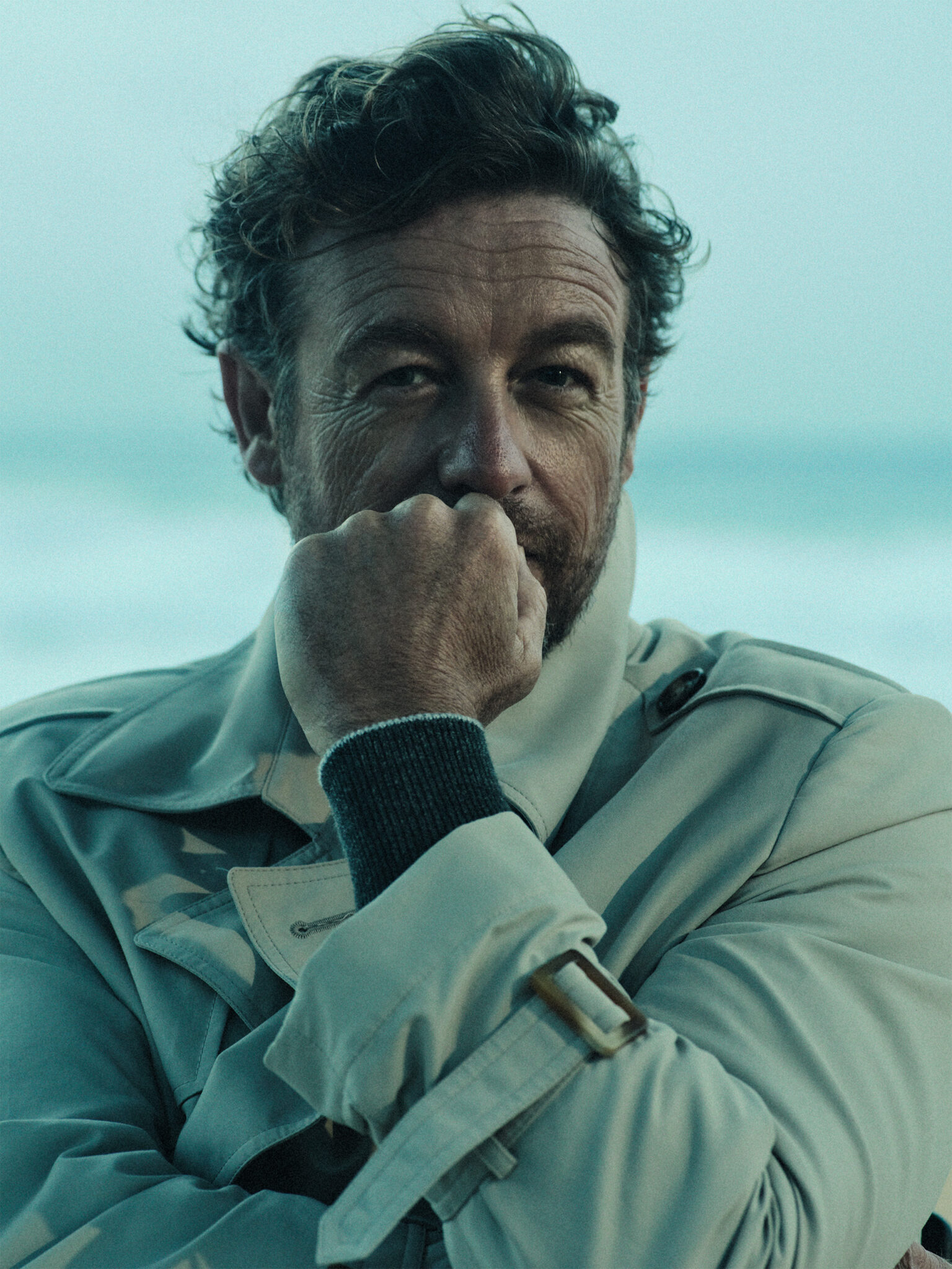 Headshot of a light-skinned adult man with wavy brown hair, a tan trench coat, and a dark sweater with a blurred sea background.