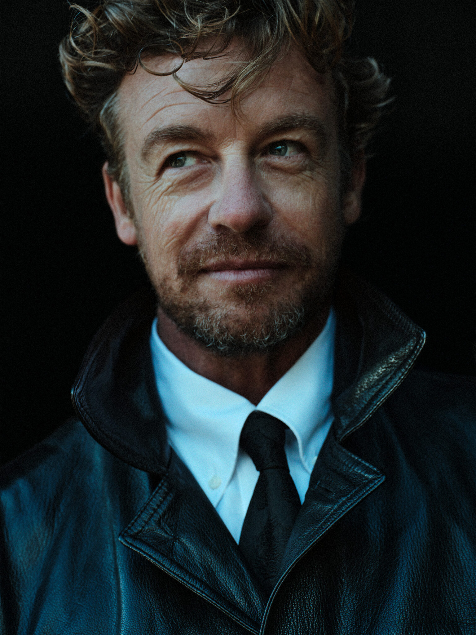 Headshot of adult male with curly hair wearing a black leather jacket, white shirt, and black tie against a dark background, looking left.