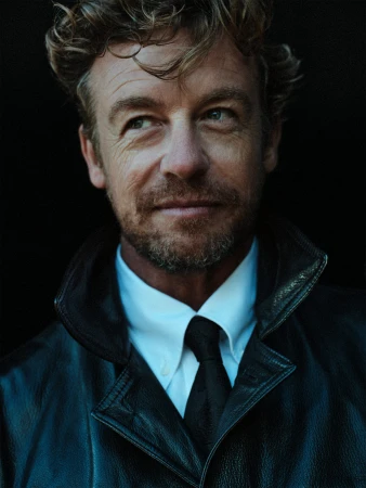 Headshot of adult male with curly hair wearing a black leather jacket, white shirt, and black tie against a dark background, looking left.