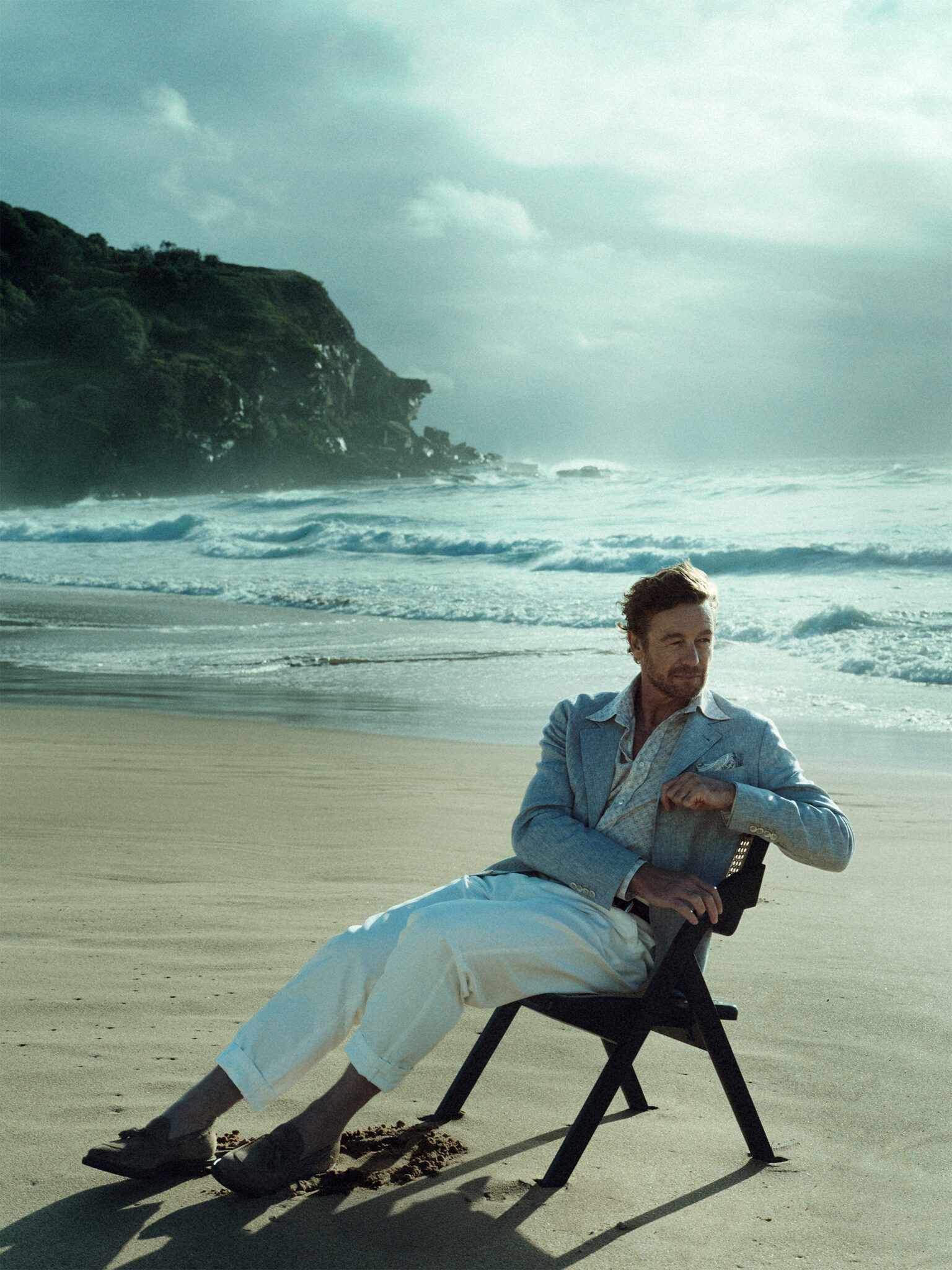 A relaxed man sits on a black folding chair on a beach with an ocean view, wearing a blazer and white pants, enjoying the calming coastal scenery.