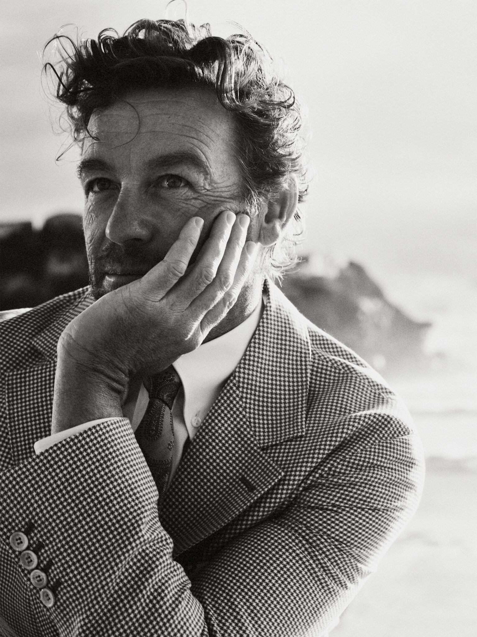 Black and white portrait of a man in a patterned blazer, hand to his face, near the coast with rocks and ocean in the background.