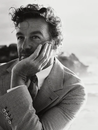 Black and white portrait of a man in a patterned blazer, hand to his face, near the coast with rocks and ocean in the background.