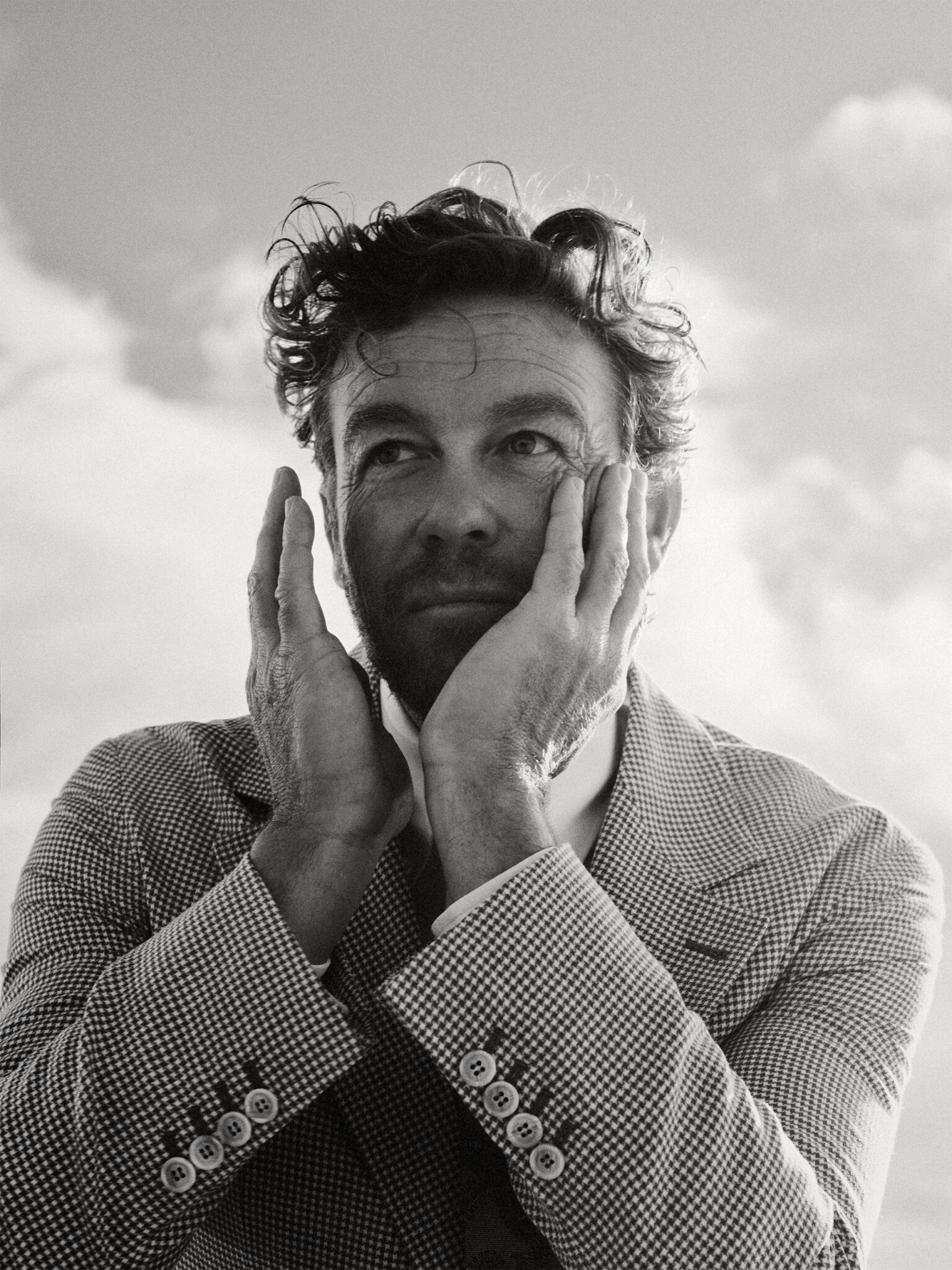 Black-and-white portrait of an adult man with textured hair, wearing a patterned blazer, holding his face with both hands against a cloudy sky.