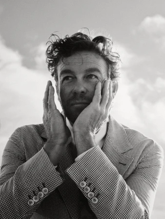 Black-and-white portrait of an adult man with textured hair, wearing a patterned blazer, holding his face with both hands against a cloudy sky.