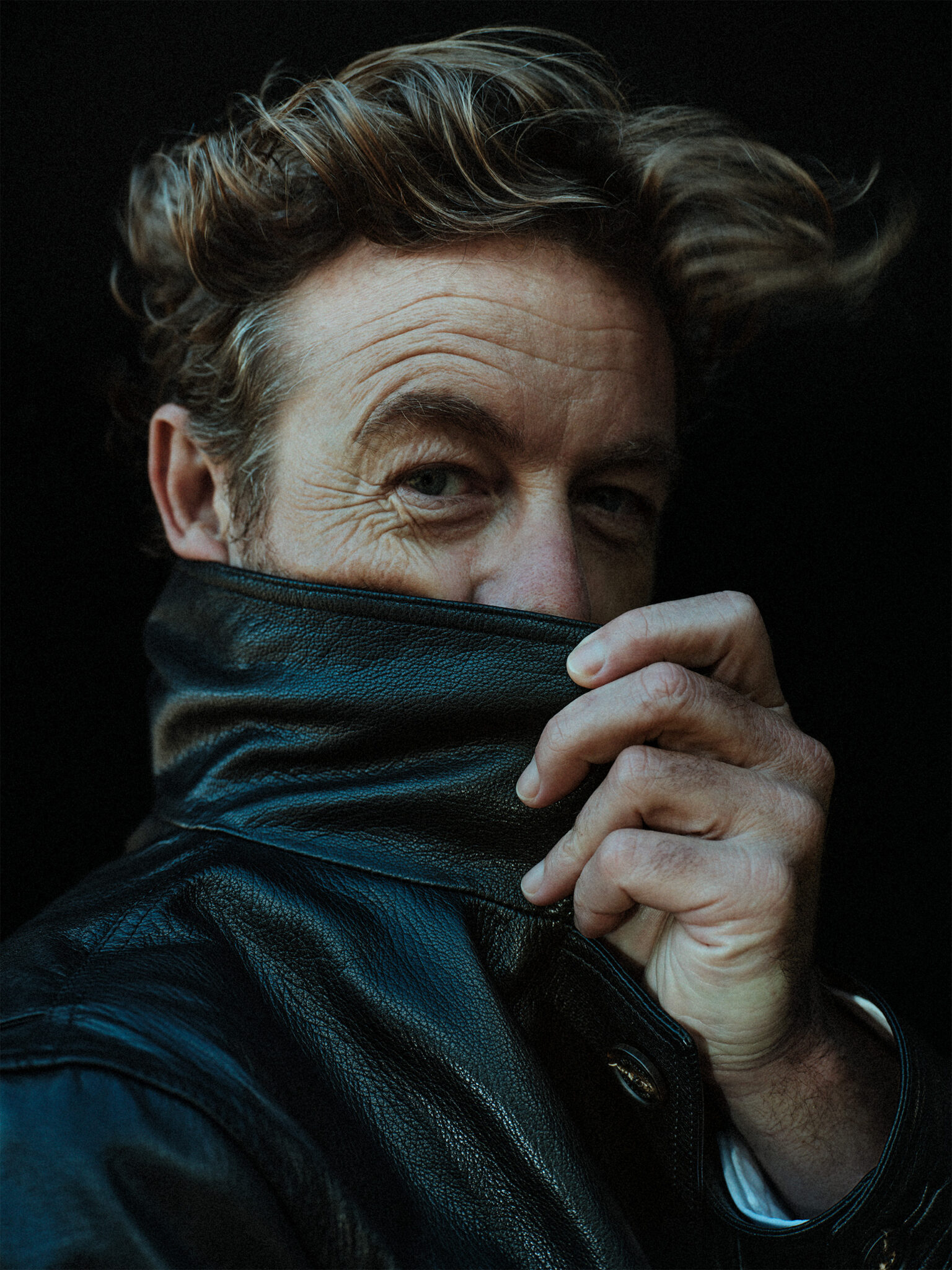 Portrait of a fair-skinned adult male in a black leather jacket, partially obscuring his face with the jacket's collar, against a dark backdrop.