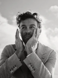 Black and white portrait of Simon Baker holding his hands against his face wearing a patterned blazer against a cloudy sky background.